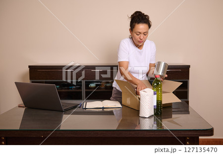 Woman Packing Food Items into a Box While Using a Laptop at Home Woman Packing Food Items into a Box While Using a Laptop at Home 127135470