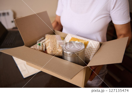 Person Holding A Cardboard Box Filled with Canned Goods and Dry Food Items 127135474