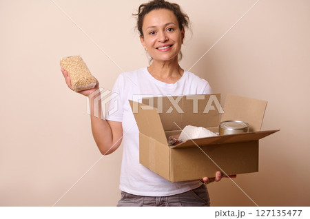 Woman Smiling While Displaying a Food Donation Package Including Grains and Canned Goods 127135477