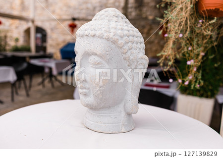 Stone Buddha head sculpture on a white table in outdoor cafe. Mindfulness, spiritual ambiance, and Asian-inspired decor. Stone Buddha head sculpture on a white table in outdoor cafe. Mindfulness, spiritual ambiance, and Asian-inspired decor. 127139829