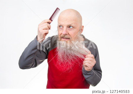 Elderly bald man with a thick white beard uses a on his head, pulling beard hair with his other hand 127140583