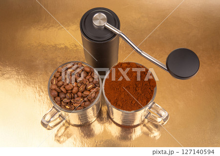 A manual coffee grinder sits between two glass mugs, one with roasted coffee beans A manual coffee grinder sits between two glass mugs, one with roasted coffee beans 127140594