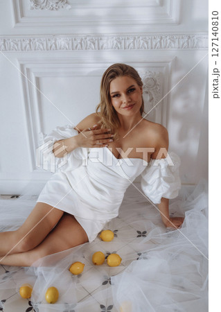 Elegant woman in white dress sitting on tiled floor in classic white room with lemons 127140810