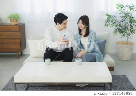 リビングでスマホを見る若い夫婦　 127142959