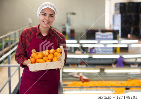 Glad positive female employee in colored uniforms hold a box of fresh ripe tangerines in their hands on citrus sorting line at warehouse. Glad positive female employee in colored uniforms hold a box of fresh ripe tangerines in their hands on citrus sorting line at warehouse. 127148203