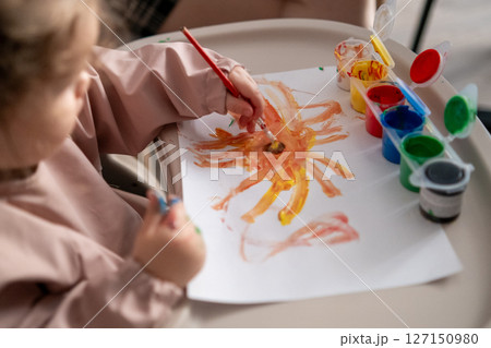 Little girl painting a bright yellow sun with gouache. Childs hand holding a paintbrush, early art skills, imagination and color exploration. Little girl painting a bright yellow sun with gouache. Childs hand holding a paintbrush, early art skills, imagination and color exploration. 127150980