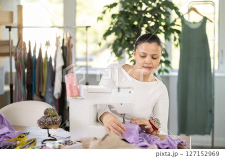 Attentive woman tailor sewing textile using sewing machine sitting at workplace in fashionable sewing atelier Attentive woman tailor sewing textile using sewing machine sitting at workplace in fashionable sewing atelier 127152969