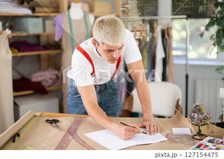 Focused young tailor drawing sketches in sewing workshop Focused young tailor drawing sketches in sewing workshop 127154475