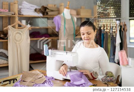 Positive young female dressmaker working with sewing-machine in tailor's workshop Positive young female dressmaker working with sewing-machine in tailor's workshop 127154538