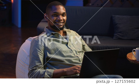 Focused black person multitasking on laptop in stylish loft space, connected to the internet while managing daily freelance tasks and remote responsibilities. Browsing web on sofa. Camera B. 127155602