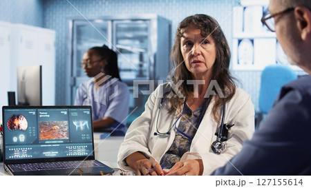 Senior doctor at a consultation with patient reads the radiography scans in cancer prevention and early diagnosis, offering personalized treatment recommendations. Risk factors. Camera A. Senior doctor at a consultation with patient reads the radiography scans in cancer prevention and early diagnosis, offering personalized treatment recommendations. Risk factors. Camera A. 127155614