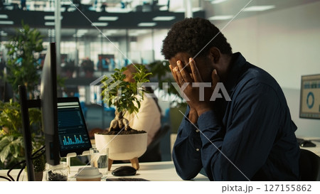 Overworked man having migraine symptoms while working on PC, drinking water to recover. Tired office worker in pain, feeling discomfort while doing tasks on PC, doing rehydration, camera B 127155862