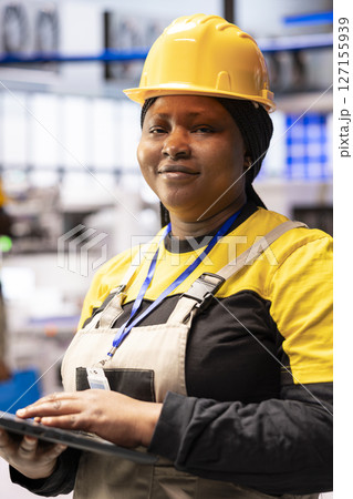 Portrait of female automation expert overseeing optimization of robot arm and assembly line in industrial plant. Closeup of african american industry 4.0 engineer in smart factory, looking at camera. Portrait of female automation expert overseeing optimization of robot arm and assembly line in industrial plant. Closeup of african american industry 4.0 engineer in smart factory, looking at camera. 127155939