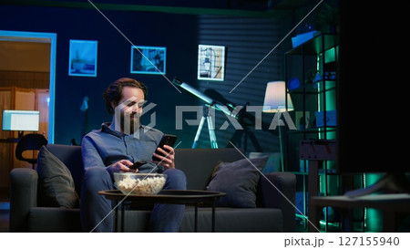 Smiling person on couch picking movie on streaming service using remote and texting on mobile phone. Happy man watching shows on television, zapping though channels, chatting on smartphone, camera B Smiling person on couch picking movie on streaming service using remote and texting on mobile phone. Happy man watching shows on television, zapping though channels, chatting on smartphone, camera B 127155940