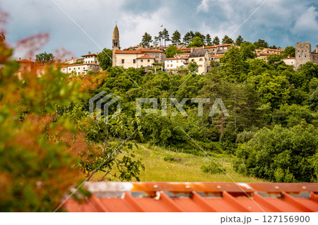 Scenic Hilltop Town Of Stanjel In Slovenia: Traditional Stone Houses And Historic Church Tower Rising Above Lush Greenery Under A Dramatic Cloudy Sky In The Karst Region 127156900