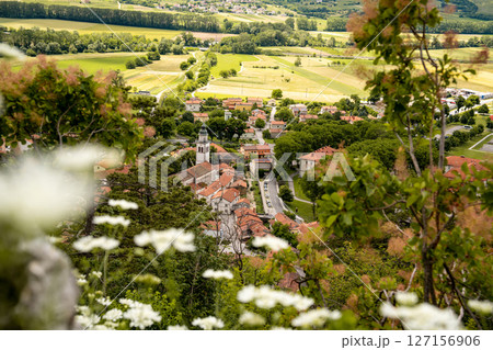 Picturesque View Of Vipava From Castle Ruins: Red-Tiled Rooftops And Church Tower Nestled In Green Valley With Blooming Flowers In Foreground And Rolling Fields In Background, Slovenia Picturesque View Of Vipava From Castle Ruins: Red-Tiled Rooftops And Church Tower Nestled In Green Valley With Blooming Flowers In Foreground And Rolling Fields In Background, Slovenia 127156906