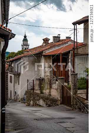 Rustic Street Scene In Vipava, Slovenia: Old Stone House With Wooden Doors And A Bench Beneath Overhanging Green Vines Along A Quiet Winding Lane In The Countryside 127156921