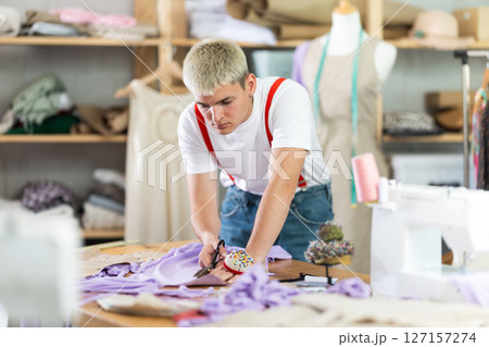 Attentive young tailor cutting textile in sewing studio 127157274