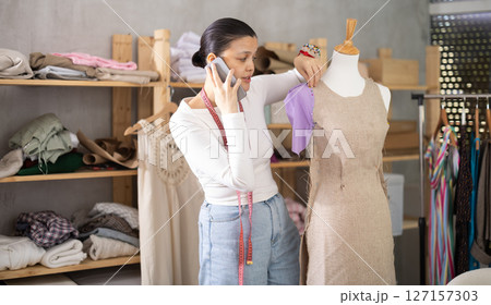Experienced young female fashion designer working with fabric and mannequin while sewing fashionable dress, consulting with client on mobile phone in sewing workshop Experienced young female fashion designer working with fabric and mannequin while sewing fashionable dress, consulting with client on mobile phone in sewing workshop 127157303