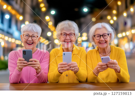 Three joyful older women engaged with their smartphones in a lively nighttime atmosphere adorned with fairy lights 127159813