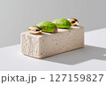 Dessert placed on clean white surface with shiny golden decorations and vibrant green toppings. Minimalist setting showcases modern food presentation 127159827