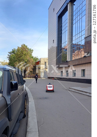 An autonomous delivery robot moves along a city street near a modern building, showcasing urban automation 127161786