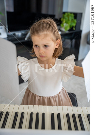Little girl reading sheet music before piano practice at home. Focused moment of musical learning and preparation in a cozy family setting. Little girl reading sheet music before piano practice at home. Focused moment of musical learning and preparation in a cozy family setting. 127161967
