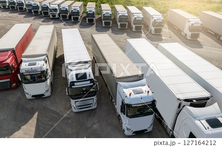 Aerial view of a large truck parking lot at a logistics hub, with multiple commercial semi trucks parked in organized rows. Aerial view of a large truck parking lot at a logistics hub, with multiple commercial semi trucks parked in organized rows. 127164212