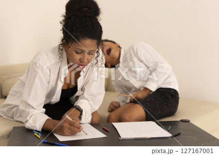 Focused woman drawing while tired boy yawns in the background Focused woman drawing while tired boy yawns in the background 127167001