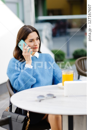 Young woman talking on smartphone and drinking orange juice at outdoor cafe 127167607