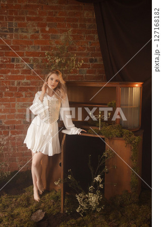 Young woman in white dress standing barefoot near vintage cabinet against red brick wall Young woman in white dress standing barefoot near vintage cabinet against red brick wall 127168182