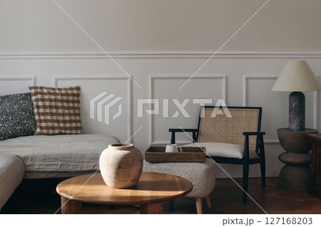 Serene cozy living room bathed in natural light. Neutral color palette. Wooden coffee table, wicker chair, tray with cup of tea coffee. Soft cushions on sofa, minimalistic decor. Empty wall bacground. Serene cozy living room bathed in natural light. Neutral color palette. Wooden coffee table, wicker chair, tray with cup of tea coffee. Soft cushions on sofa, minimalistic decor. Empty wall bacground. 127168203