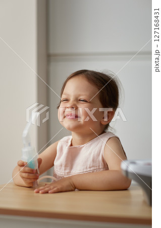 cute little toddler girl holds nebulizer in her hand and smiles sitting at table cute little toddler girl holds nebulizer in her hand and smiles sitting at table 127168431