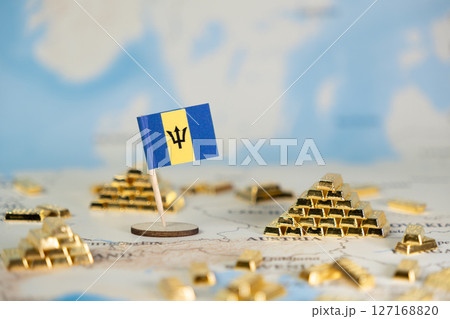 Miniature Barbados flag standing alongside piles of shiny gold bars on a blurred world map, symbolizing national wealth, economic stability, and global investment 127168820