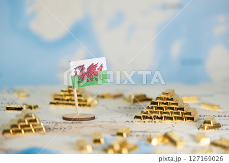 Welsh national flag standing prominently among gleaming gold bar pyramids and scattered assets on a blurred global map, conceptualizing financial prosperity and economic independence for the nation 127169026