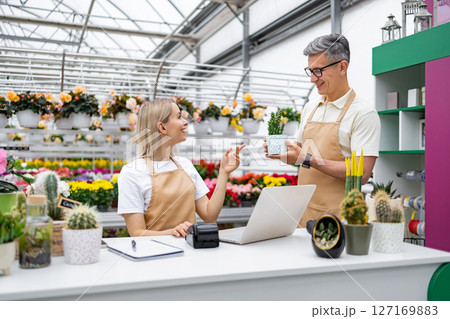 A Caucasian man and woman are in a flower shop, interacting with each other while looking at a small plant. 127169883