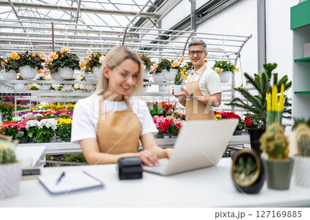 A Caucasian florist and seller happily working at their shop's checkout. Plants fill the background, and a man poses with a potted plant. 127169885