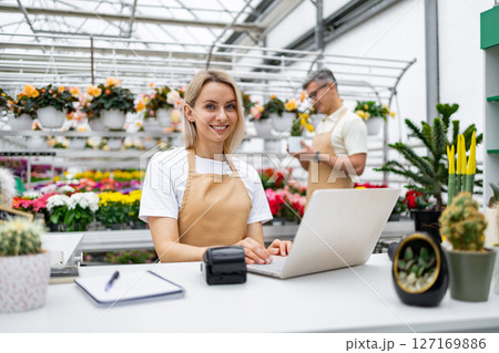 A florist is at work, smiling and using a laptop. In the background, a colleague is inspecting a plant, in a sunny greenhouse filled with flowers. 127169886