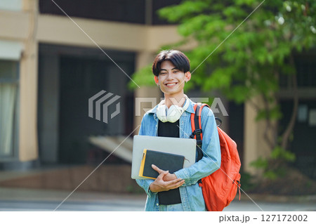 A cheerful Asian college student stands outside on campus with a backpack, smiling and looking at the camera. 127172002