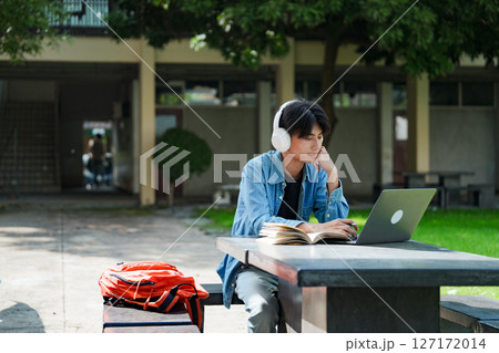 Portrait of a young Asian male college student e-learning using laptop on the bench on campus. 127172014