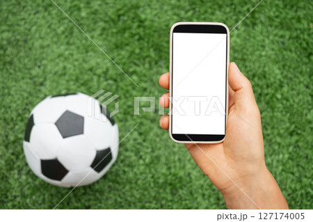 Man hand holds smartphone with white blank screen next to soccer ball on green grass. Online. Modern technology with traditional sports. Space for text. View from above. Empty display . Man hand holds smartphone with white blank screen next to soccer ball on green grass. Online. Modern technology with traditional sports. Space for text. View from above. Empty display . 127174005