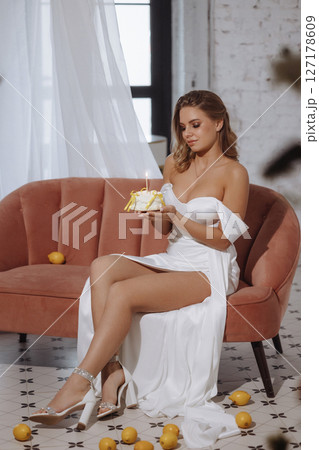 Woman in white dress sitting on pink sofa holding birthday cake with candle in classic room Woman in white dress sitting on pink sofa holding birthday cake with candle in classic room 127178609