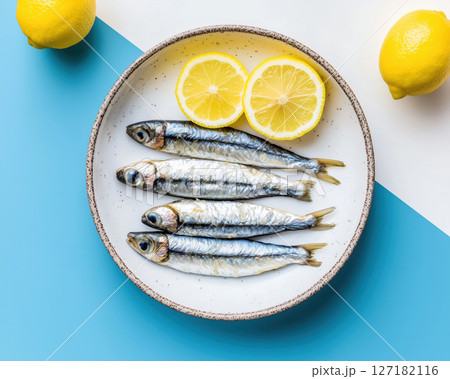 A blue plate with fresh sardines, lemon slices, and sea salt on a vibrant blue and white background. 127182116
