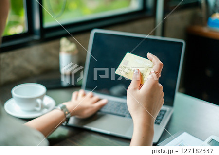 A woman uses a laptop and credit card for online shopping in a cozy cafe, with daylight streaming through the window. A woman uses a laptop and credit card for online shopping in a cozy cafe, with daylight streaming through the window. 127182337