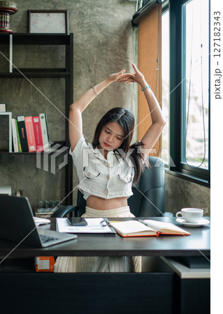 A young woman takes a break to stretch at her office desk, promoting wellness and productivity in a modern workspace. A young woman takes a break to stretch at her office desk, promoting wellness and productivity in a modern workspace. 127182343