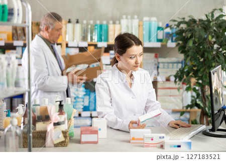 Girl pharmacist uses computer at workplace. Male colleague in background 127183521