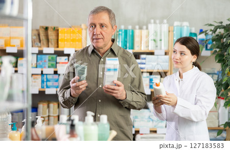 Senior man buys cream at drug store, girl pharmacist in background Senior man buys cream at drug store, girl pharmacist in background 127183583