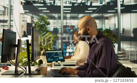 Accountant in busy workplace talking with colleagues while inputting financial figures on PC. Employee working in modern office chatting with coworkers while crosschecking data, camera B 127183658