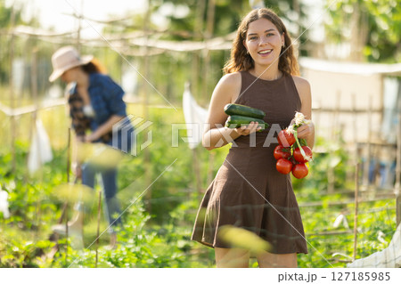 Positive woman in dress posing with tomatoes, zucchini and green onions in garden 127185985