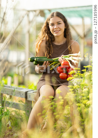 Positive woman in dress posing with tomatoes, zucchini and green onions in garden 127185996